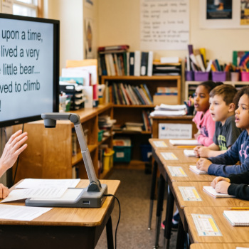 A teacher leads an echo reading lesson, gesturing as students read aloud together from a shared text projected on a large screen displaying the words "Once upon a time, there lived a very brave little bear... He loved to climb trees!"