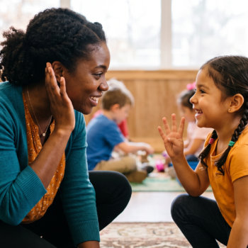 A teacher kneels eye-to-eye with a young girl who holds up four fingers, counting sounds together, while other students play in the background of a sunlit classroom.