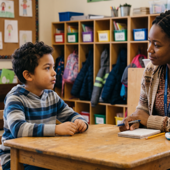 A teacher with a notepad and pen sits across from a young boy at a small table, listening attentively as he responds during a one-on-one phonological awareness assessment.