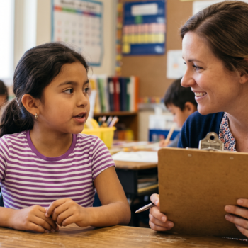 A teacher sits beside a young elementary student at a classroom desk, smiling warmly as the child speaks. The teacher holds a clipboard and pen, suggesting an informal phonemic awareness assessment in progress. Other students work independently in the background.