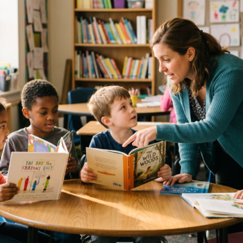 A teacher leans over a small table, guiding four young readers through a discussion of their picture books.
