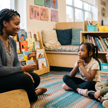A teacher sits in a low chair facing two students on a classroom rug, smiling warmly as the children listen attentively during a small group onset-rime awareness lesson.