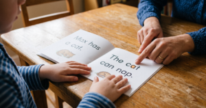 A teacher points to words in a decodable reader showing the sentences "Max has a cat" and "The cat can nap" while a young child follows along with both hands on the page.