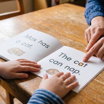 A teacher points to words in a decodable reader showing the sentences "Max has a cat" and "The cat can nap" while a young child follows along with both hands on the page.