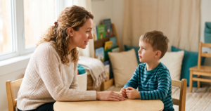 A teacher leans in to talk with a young student at a small table in a cozy reading corner