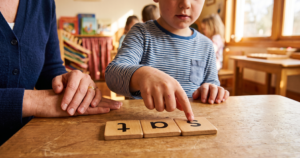 A young child points to wooden letter tiles spelling "sat" on a table during a synthetic phonics lesson, with a teacher seated alongside providing support.