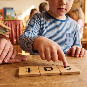 A young child points to wooden letter tiles spelling "sat" on a table during a synthetic phonics lesson, with a teacher seated alongside providing support.