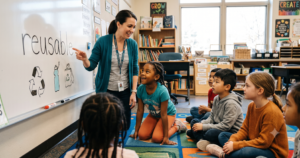 Teacher pointing to the word "reusable" on a whiteboard while students seated on a carpet engage in a base words lesson in an elementary classroom