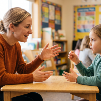 Teacher sitting face-to-face with a young student using hand gestures to teach phonological awareness at a small classroom table