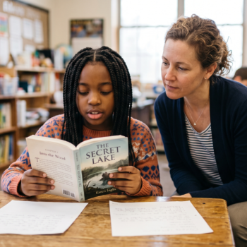 Teacher listening closely as an elementary student reads aloud from a chapter book at her desk in a classroom library setting